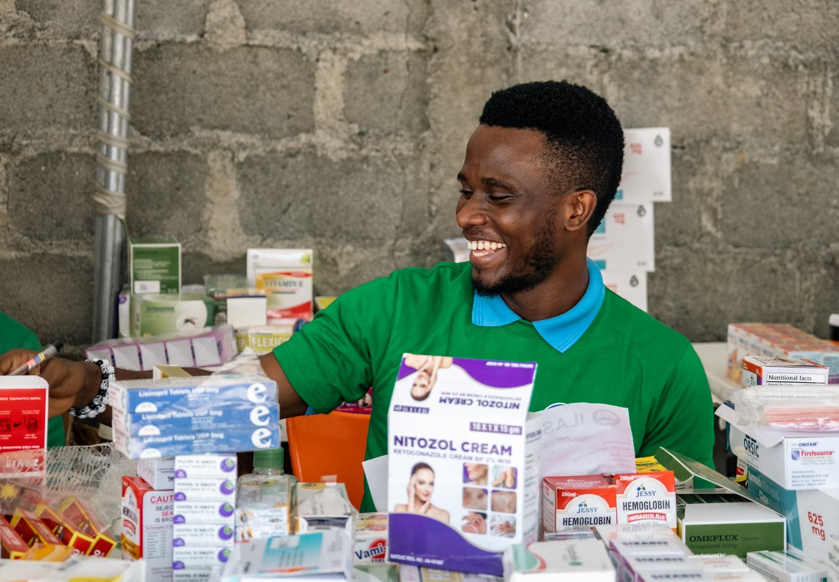 A volunteer pharmacist smiling as he works behind a table stacked with boxed medicines at the Ilasan medical outreach in Lekki, Lagos.