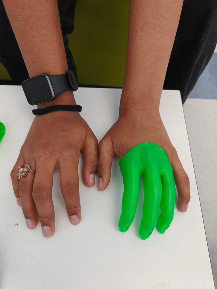 A hand resting on a table beside a green 3D printed prosthetic hand.