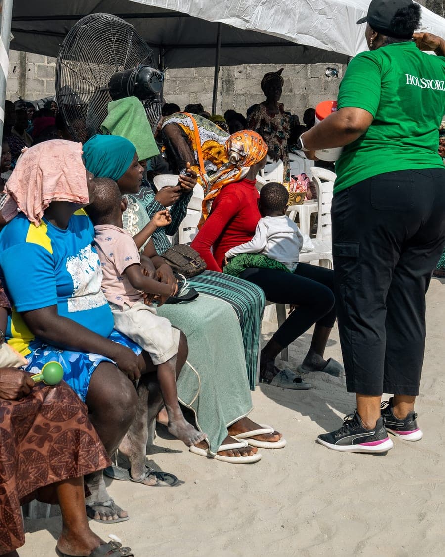 A volunteer in a Foundation tee stands addressing a seated audience under the marquee at the Ilasan medical outreach in Lekki, Lagos.