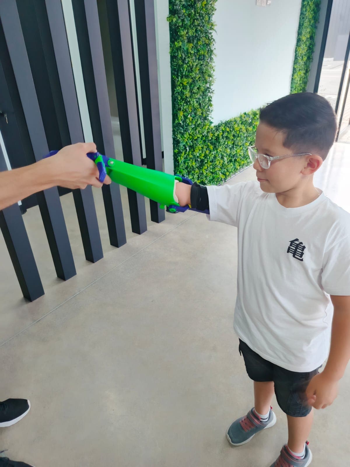 A boy in glasses reaching out as an adult fits a green and blue 3D printed arm to him.