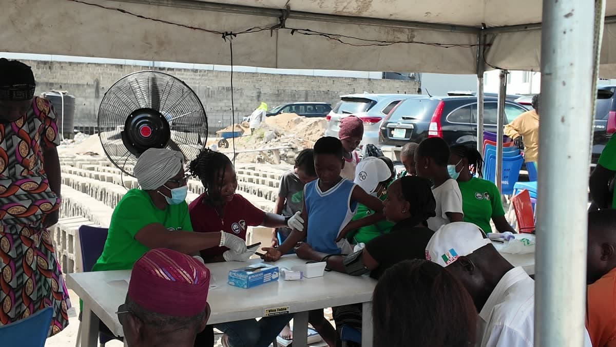 A wide view under the marquee at the Ilasan medical outreach in Lekki, Lagos, with volunteers working and people waiting on rows of chairs.